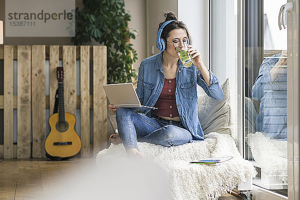 Frau mit Kopfhörern und Laptop sitzt zu Hause am Fenster und trinkt aus Glas
