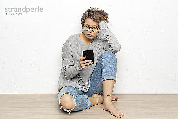 Porträt einer jungen Frau mit drahtlosen Kopfhörern  die auf dem Boden sitzt und auf ihr Handy schaut