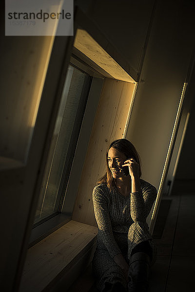 Frau sitzt bei Sonnenuntergang am Bürofenster und telefoniert