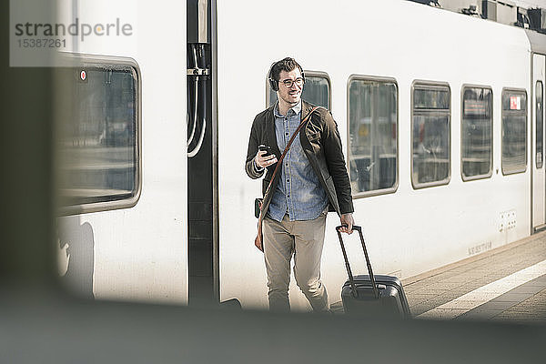 Lächelnder junger Mann mit Kopfhörern  Handy und Koffer auf dem Bahnsteig