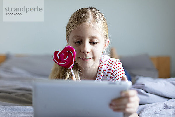 Porträt eines blonden  auf dem Bett liegenden Mädchens mit Lolli mit digitalem Tablett