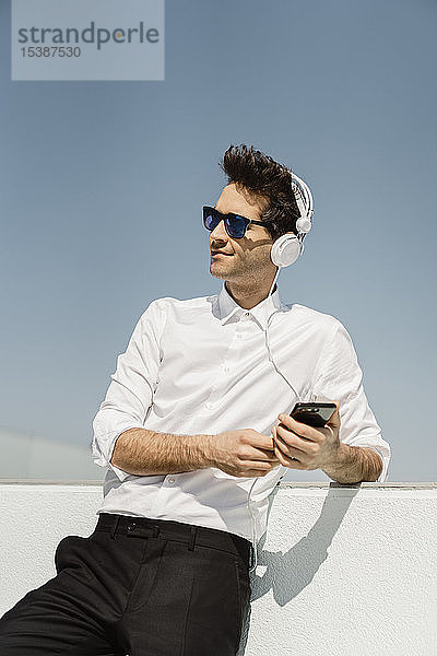 Porträt eines Geschäftsmannes  der auf der Dachterrasse mit Kopfhörern und Smartphone Musik hört