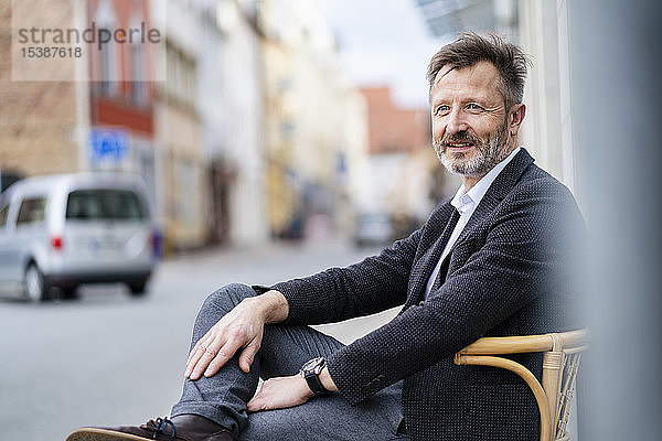 Porträt eines zufriedenen reifen Geschäftsmannes mit grauem Bart auf einem Stuhl in der Stadt sitzend