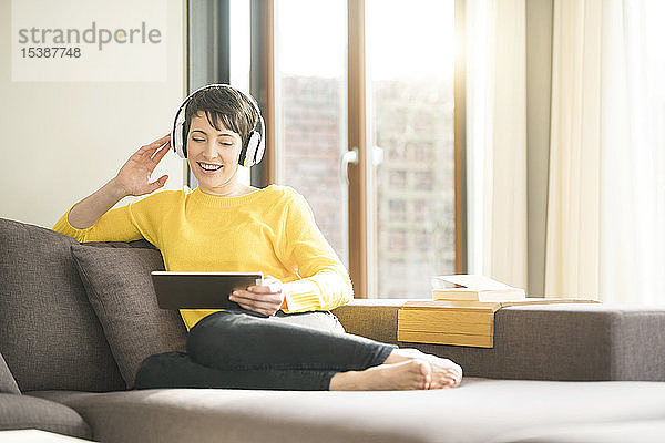 Porträt einer glücklichen Frau  die zu Hause auf der Couch sitzt und mit Kopfhörern Musik hört  während sie auf das Tablett schaut