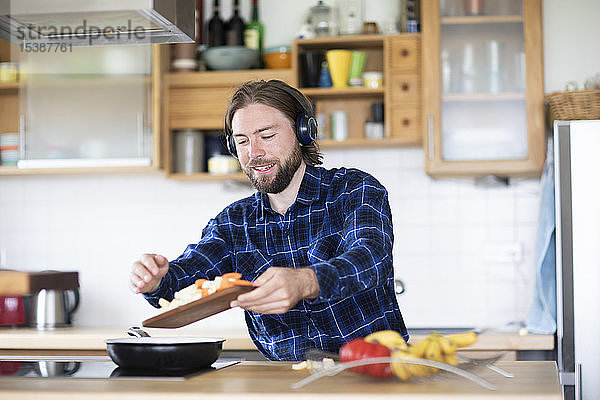 Junger Mann mit Bart und kariertem Hemd und Kopfhörer  der in der Küche Gemüse kocht