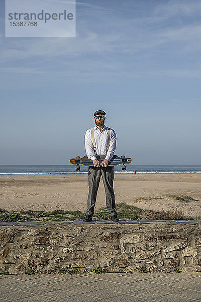 Mann mit Longboard steht auf einer Mauer vor Strand und Meer