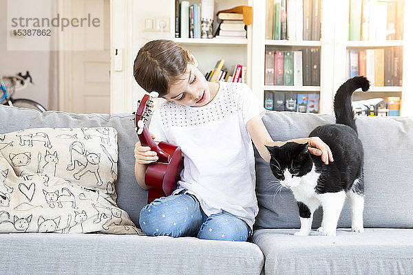 Mädchen mit Ukulele sitzt auf Couch und streichelt Katze