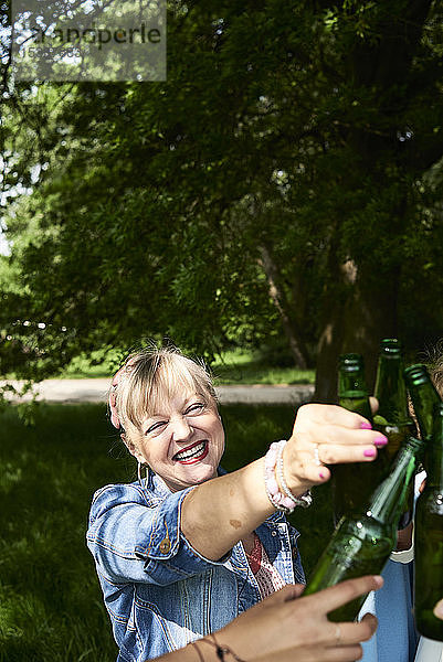 Porträt einer glücklichen Frau  die mit Freunden im Park an einer Bierflasche klimpert