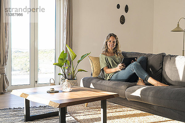 Porträt einer lächelnden Frau  die sich zu Hause auf der Couch mit Tablette vergnügt
