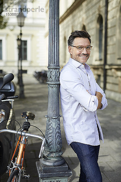 Porträt eines lächelnden Geschäftsmannes mit Fahrrad  der sich in der Stadt an einen Laternenmast lehnt