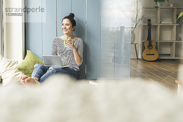 Lächelnde Frau sitzt zu Hause am Fenster  arbeitet mit Laptop und trinkt Infusionswasser