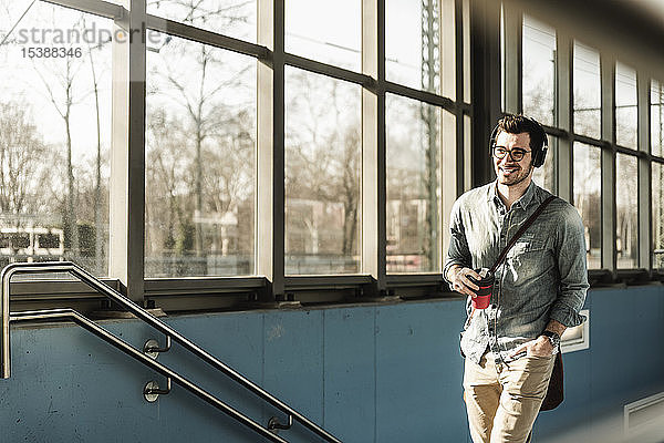 Lächelnder junger Mann mit Kopfhörern und Kaffee zum Mitnehmen auf dem Bahnhof