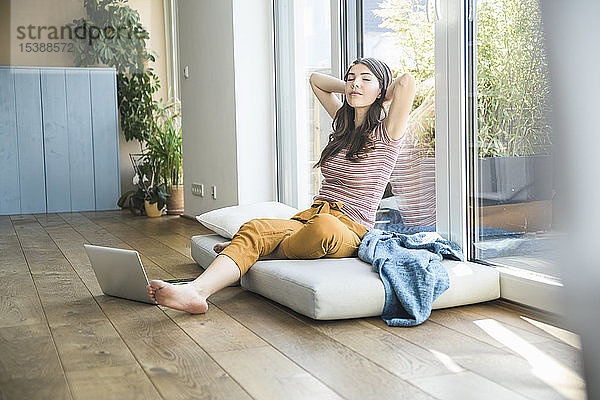 Entspannte junge Frau sitzt zu Hause mit Laptop am Fenster