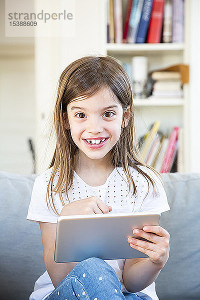 Portrait eines glücklichen kleinen Mädchens  das zu Hause auf der Couch sitzt und ein digitales Tablet benutzt