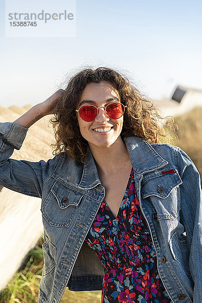 Porträt einer glücklichen jungen Frau mit lockigem braunen Haar und roter Sonnenbrille  die an einer Wand lehnt