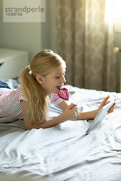 Blondes Mädchen liegt im Bett und leckt Lolli  während sie ein digitales Tablett benutzt