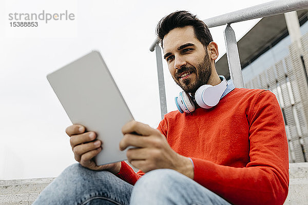 Lässiger Geschäftsmann  der auf einer Treppe in der Stadt sitzt und sein digitales Tablett und seine Kopfhörer benutzt