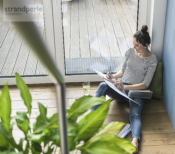 Vogelperspektive einer lächelnden Frau  die zu Hause am Fenster sitzt und mit Laptop und Aktenordner arbeitet