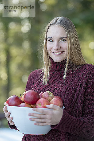 Porträt einer lächelnden blonden jungen Frau  die eine Schale mit Äpfeln hält