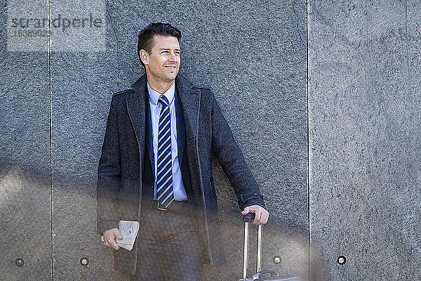 Lächelnder Geschäftsmann mit an einer Wand stehendem Koffer