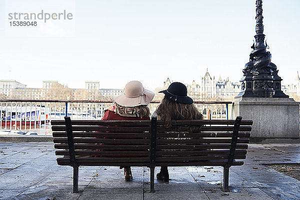 UK  London  Rückansicht von zwei Frauen  die auf einer Bank an der Uferpromenade der Themse sitzen
