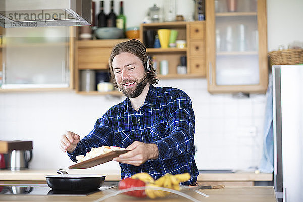 Junger Mann mit Bart und kariertem Hemd und Headset  der in der Küche Gemüse kocht