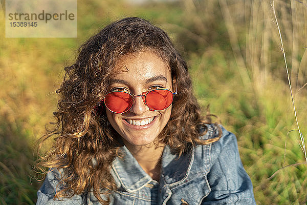 Porträt einer glücklichen jungen Frau mit lockigem braunen Haar und roter Sonnenbrille