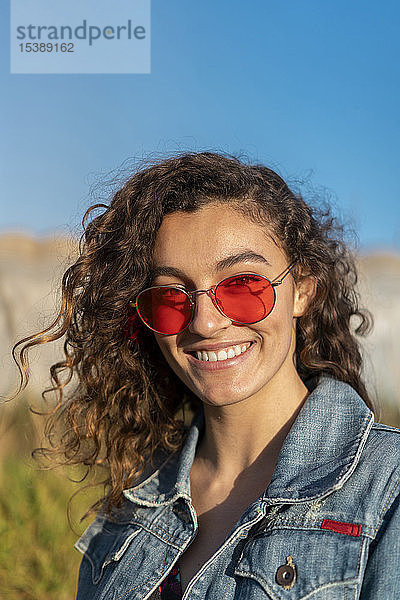 Porträt einer glücklichen jungen Frau mit lockigem braunen Haar und roter Sonnenbrille
