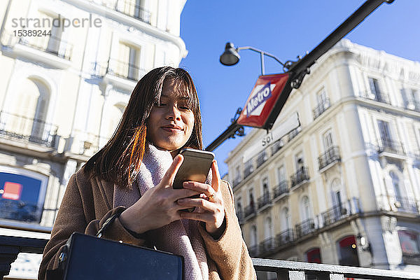 Spanien  Madrid  lächelnde junge Frau an der U-Bahn-Station Puerta del Sol beim Überprüfen ihres Smartphones