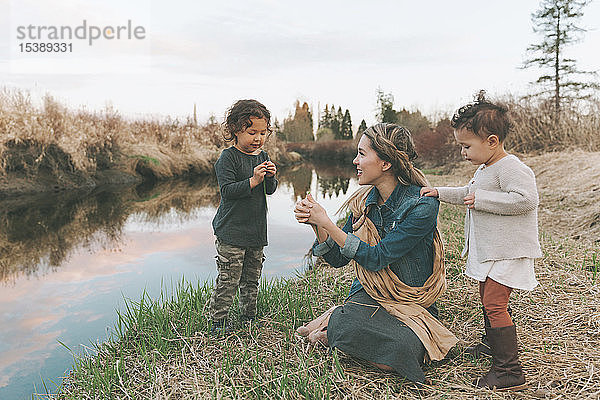 Mutter und Kinder spielen mit einem Grashalm an einem Fluss