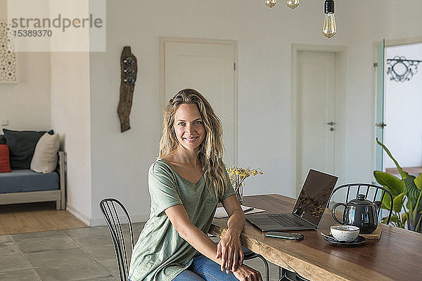Porträt einer lächelnden Frau mit Laptop  die zu Hause am Esstisch sitzt