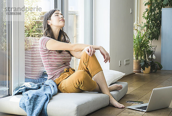 Entspannte junge Frau sitzt zu Hause mit Laptop am Fenster