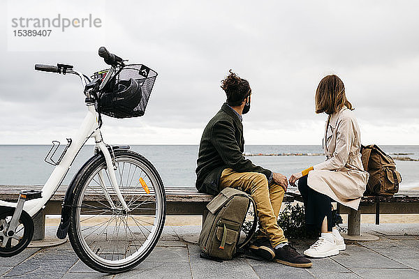 Ehepaar sitzt auf einer Bank an der Strandpromenade neben dem E-Bike und schaut auf das Meer
