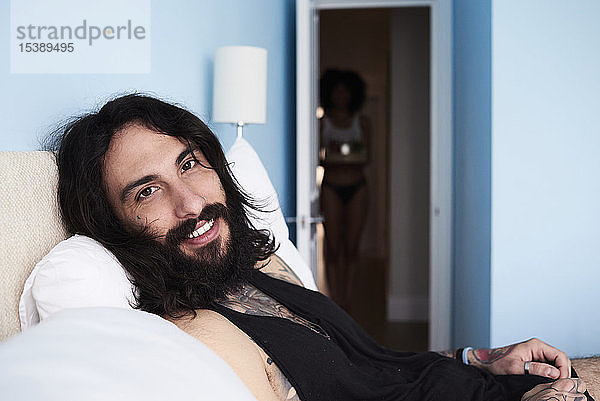 Porträt eines lächelnden tätowierten Mannes im Bett liegend mit Freundin im Hintergrund