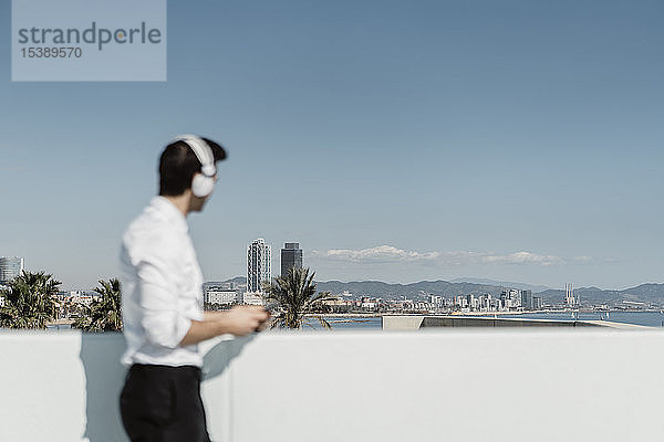 Spanien  Barcelona  Geschäftsmann hört Musik mit Kopfhörern und Smartphone auf der Dachterrasse und schaut in die Ferne