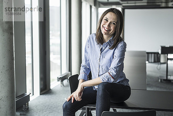 Porträt einer lächelnden Geschäftsfrau  die im Büro auf einem Konferenztisch sitzt