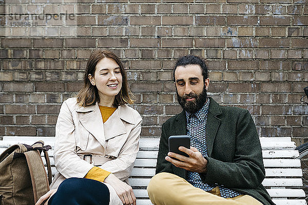 Ehepaar sitzt auf einer Bank an einer Ziegelmauer und teilt sich ein Mobiltelefon