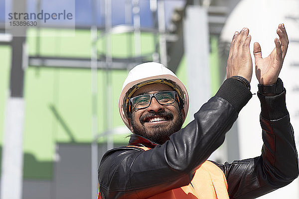 Porträt eines glücklichen Bauingenieurs vor dem Kraftwerk mit Schutzhelm und Sicherheitsweste klatschend in die Hände