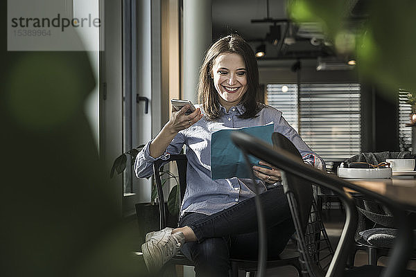 Lächelnde Geschäftsfrau sitzt im Büro mit Papier und Handy