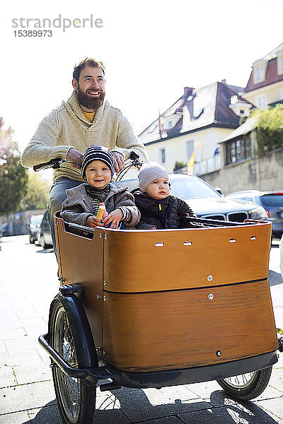 Glücklicher Vater mit zwei Kindern auf dem Lastenfahrrad in der Stadt