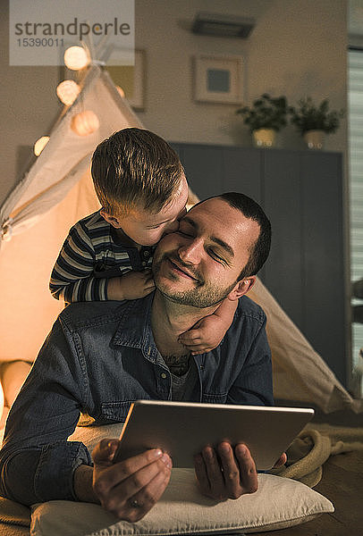 Sohn küsst Vater mit Tablette in einem beleuchteten Zelt zu Hause