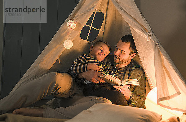 Vater liest dem Sohn in einem beleuchteten Zelt zu Hause ein Buch vor