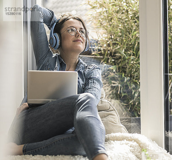 Frau mit Kopfhörern und Laptop  die zu Hause am Fenster sitzt