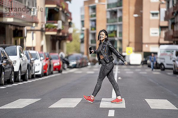 Junge Frau  die den Fußgängerüberweg überquert und ein Mobiltelefon benutzt