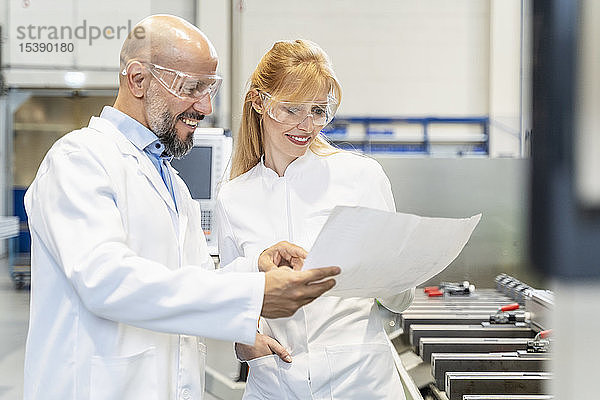 Zwei glückliche Techniker mit Laborkittel und Schutzbrille sehen sich den Plan in der Fabrik an