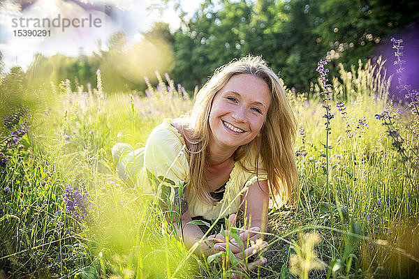 Porträt einer glücklichen blonden Frau  die im Sommer auf einer Blumenwiese liegt