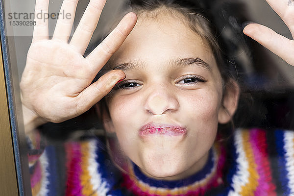Porträt eines fröhlich-verspielten Mädchens in gestreiftem Pullover hinter Fensterscheibe