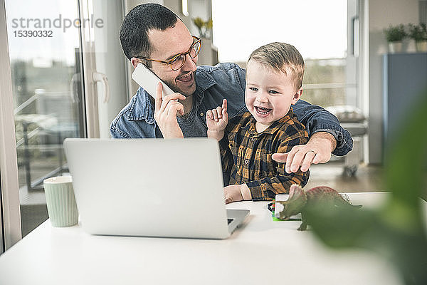 Glücklicher Vater am Handy im Heimbüro mit dem Sohn auf dem Schoß