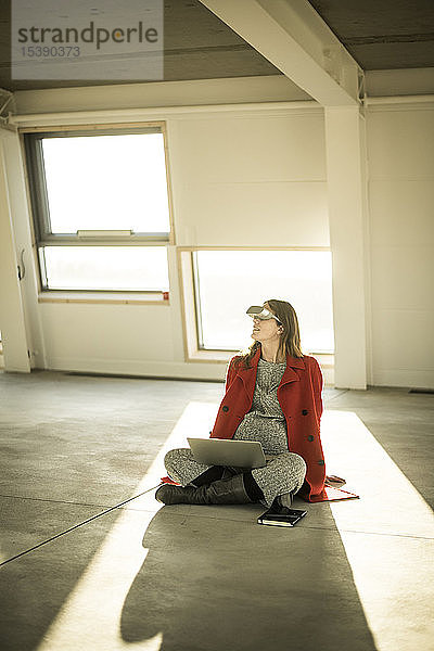 Schwangere Businessfrau sitzt auf dem Boden neuer Büroräume  benutzt VR-Brille und Laptop