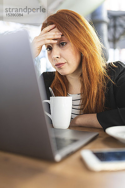 Porträt einer rothaarigen jungen Frau  die im Straßencafé auf den Laptop schaut